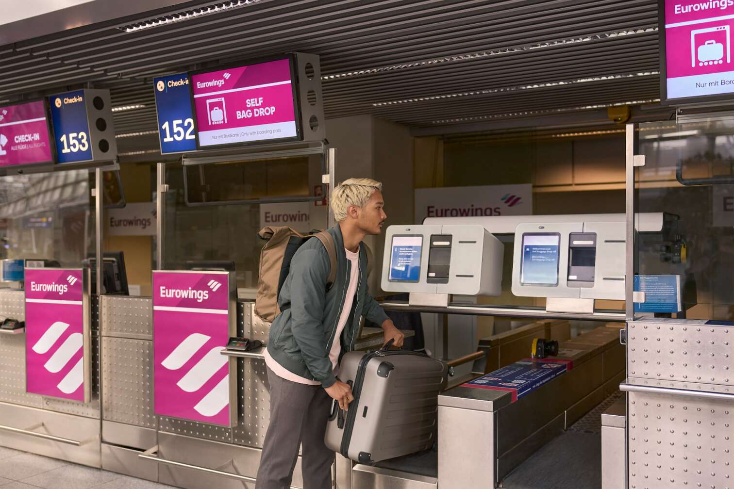 Ein junger Mann steht in einem Flughafen am Self Bag Drop von Eurowings und stellt seinen Koffer auf das Band