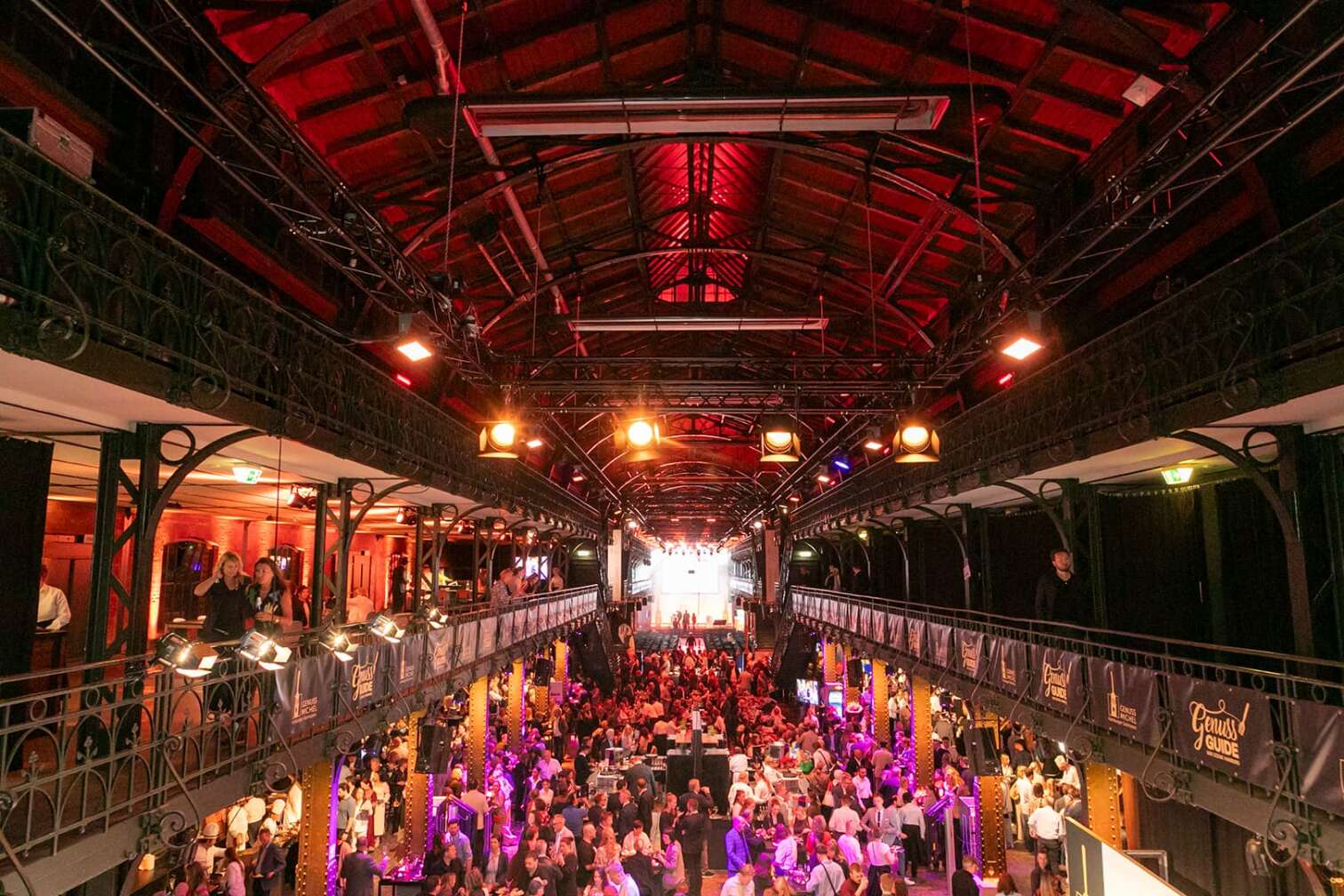 Blick in die Fischauktionshalle Hamburg während eines Genuss-Events: Zahlreiche Besucher stehen und sitzen an Tischen, probieren Speisen und Getränke. Die Halle ist festlich beleuchtet mit roten und gelben Lichtern, Banner mit der Aufschrift ‚Genuss Guide‘ hängen an den Galerien. Kulinarisches Festival, Food-Event und gesellschaftliches Treffen in Hamburg.