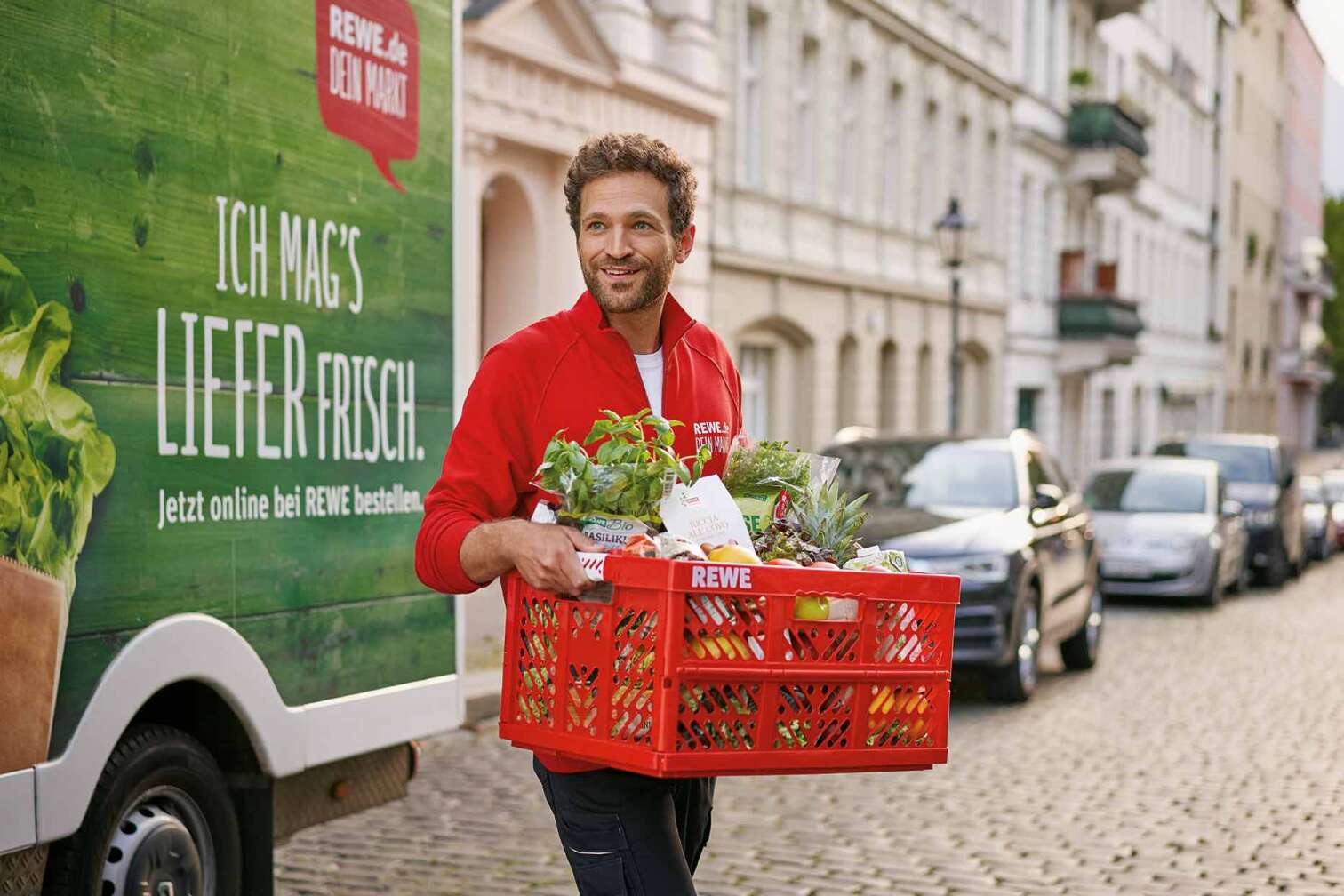 Bild eines gut gelaunten REWE Lieferservice Mitarbeiters, der eine Kiste frischer Lebensmittel trägt