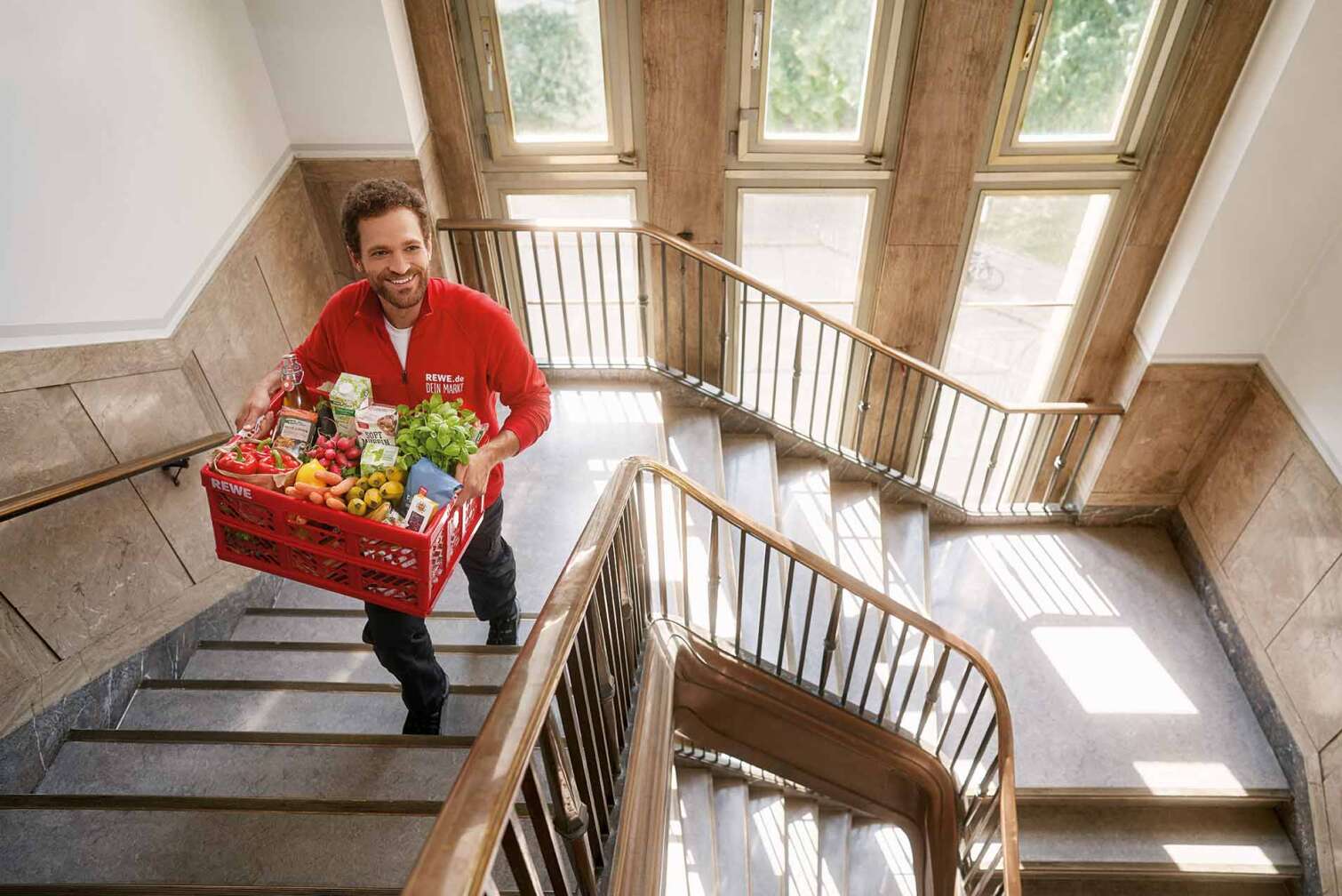 Bild eines REWE Lieferservice Mitarbeiters, der eine Kiste frischer Lebensmittel ein Treppenhaus hinauf trägt