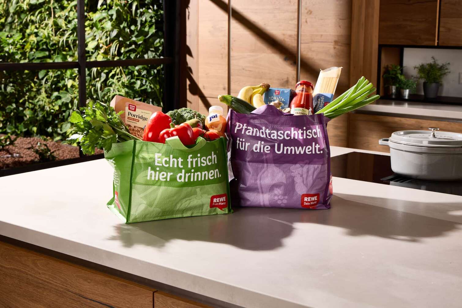 Die neuen Pfand-Mehrwegtaschen des REWE Lieferservice mit frischen, regionalen Produkten gefüllt, stehen auf einer Küchenablage