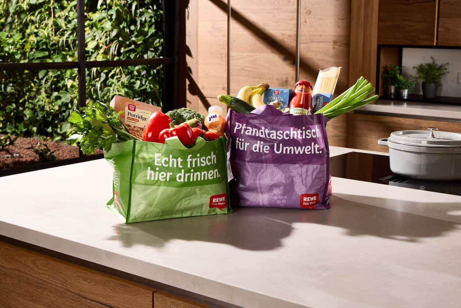 Die neuen Pfand-Mehrwegtaschen des REWE Lieferservice mit frischen, regionalen Produkten gefüllt, stehen auf einer Küchenablage